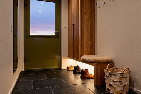 A Scandinavian-style mudroom features a tall slatted oak cabinet with an integrated bench and round beige wool cushion, illuminated by an LED strip beneath. The floor is laid in charcoal slate tiles, the walls are warm beige, and an olive green metal door with a glazed upper panel shows the dusk sky. Minimal brushed-steel hooks line the wall, with a birch-bark basket and boots arranged neatly on the floor.