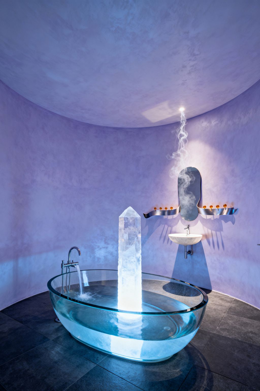 Surreal bathroom with curved lavender plaster walls and a domed ceiling, where an oval clear-glass bathtub holds a glowing crystal-like obelisk rising from its center while a freestanding chrome filler pours water at one side. Opposite, a small wall-hung stone sink sits beneath an irregular mirror flanked by wavy stainless shelves lined with amber glass bottles as a wisp of vapor drifts toward a ceiling spotlight over dark charcoal tile.