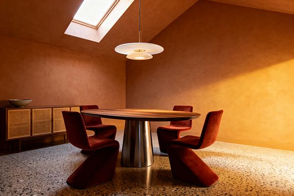 A contemporary dining room with terracotta plaster walls and a skylight above. A round walnut-top table with a brushed metal conical pedestal is encircled by four rust-colored velvet cantilever chairs. A saucer-shaped pendant hangs centrally, casting warm light. Along the left wall sits a low wood sideboard with woven cane doors and a ceramic bowl. The floor is speckled terrazzo, and sunlight from the skylight creates a soft gradient across the walls and floor.