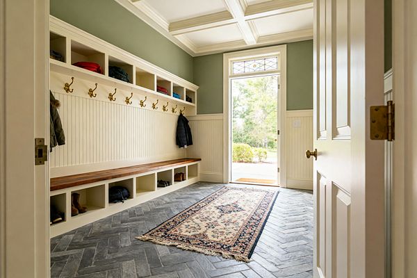 A traditional mudroom with cream beadboard wainscoting and built-in cubbies along one wall features a long walnut-stained bench and rows of brass coat hooks. Sage green upper walls, a coffered cream ceiling, and a glass exterior door with a leaded transom brighten a gray herringbone brick floor topped by a floral runner; coats, boots, and bags are neatly stored.