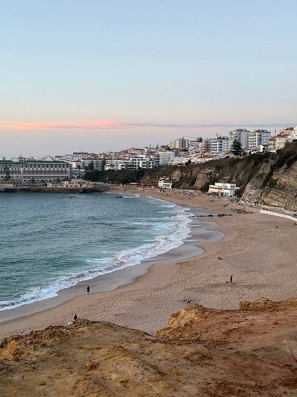 A scenic view of a beach with gentle waves and a coastal town in the background during sunset.