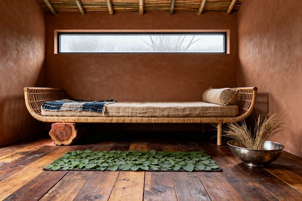 An intimate Wabi-Sabi living room features terracotta clay-plastered walls, a bamboo pole ceiling, and a long, narrow frosted window above a woven rattan daybed with a linen mattress, bolster, and frayed indigo throw. Reclaimed wood floors hold a small leaf-strewn mat, a raw tree-stump support under the daybed, and a silvery hammered metal bowl filled with dried grasses, creating a simple, earthy atmosphere.