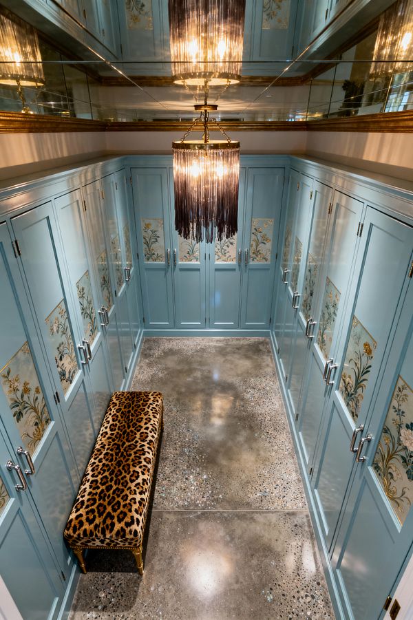 A narrow mudroom lined with powder-blue built-in cabinets features inset floral panels edged in gold and fitted with silver-tone pulls. A tiered brass chandelier with dark fringe hangs beneath mirrored ceiling panels, reflecting warm light onto a polished concrete floor speckled with aggregate. A leopard-print upholstered bench with gold legs sits along one wall, adding bold pattern to the refined cabinetry and glossy finishes.