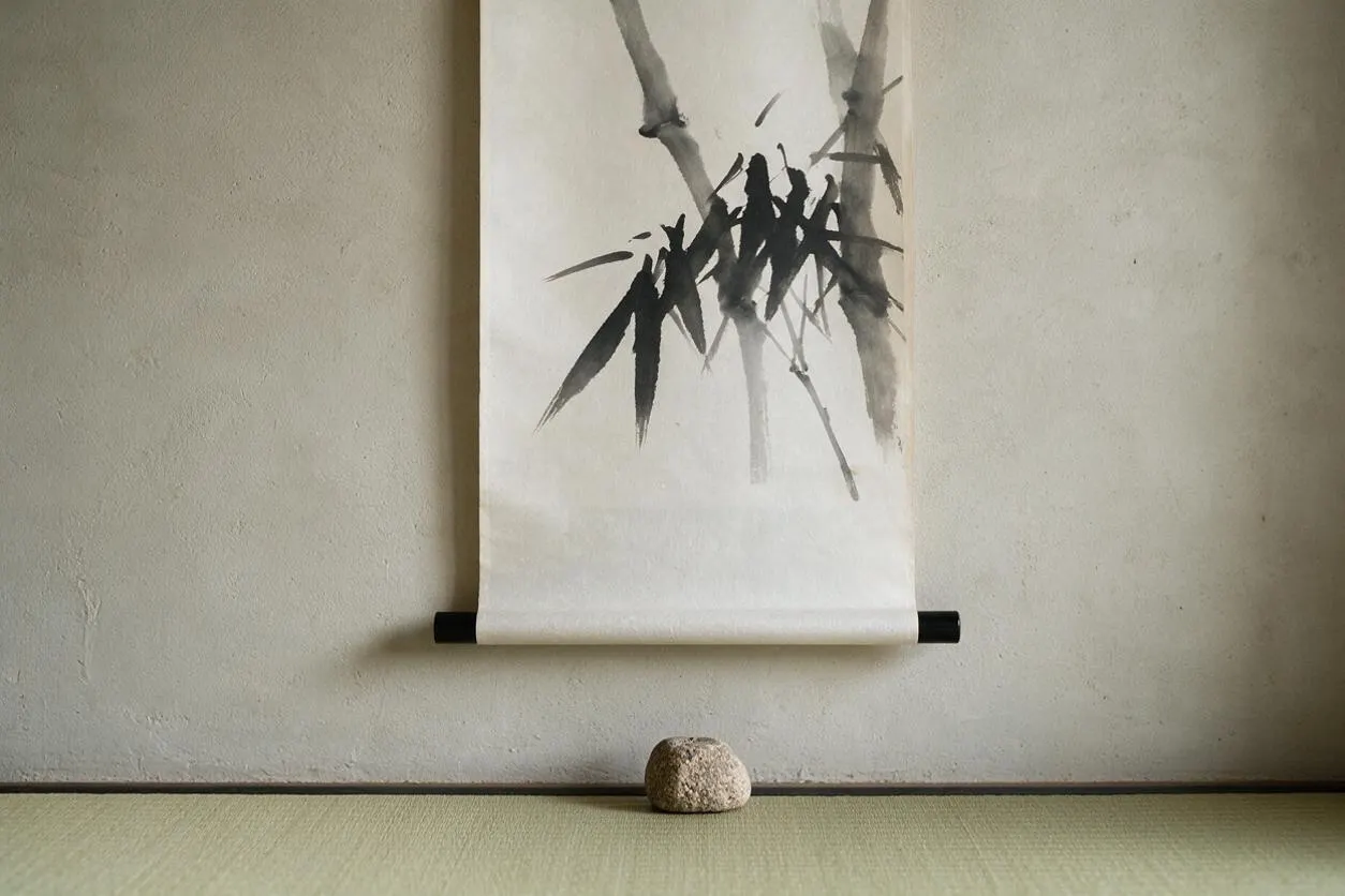 A minimalist Japanese Zen interior scene with a hanging ink scroll featuring expressive black bamboo brushwork on white paper. The scroll is mounted against a textured beige wall above a simple rounded stone placed on a pale tatami mat floor.