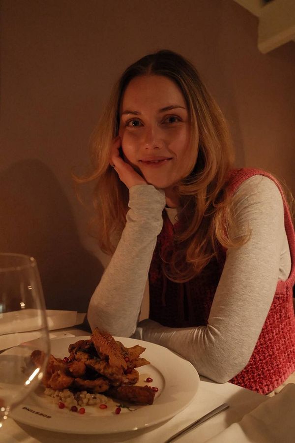 A woman is seated at a restaurant table with a plate of food in front of her, smiling warmly.