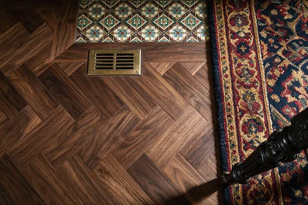 A close-up view of a Victorian-style interior floor featuring rich herringbone wood planks, a brass floor vent, a strip of decorative patterned tile, and the edge of a traditional Persian-style rug. Dark carved furniture legs appear at the lower right, adding to the classic period feel.