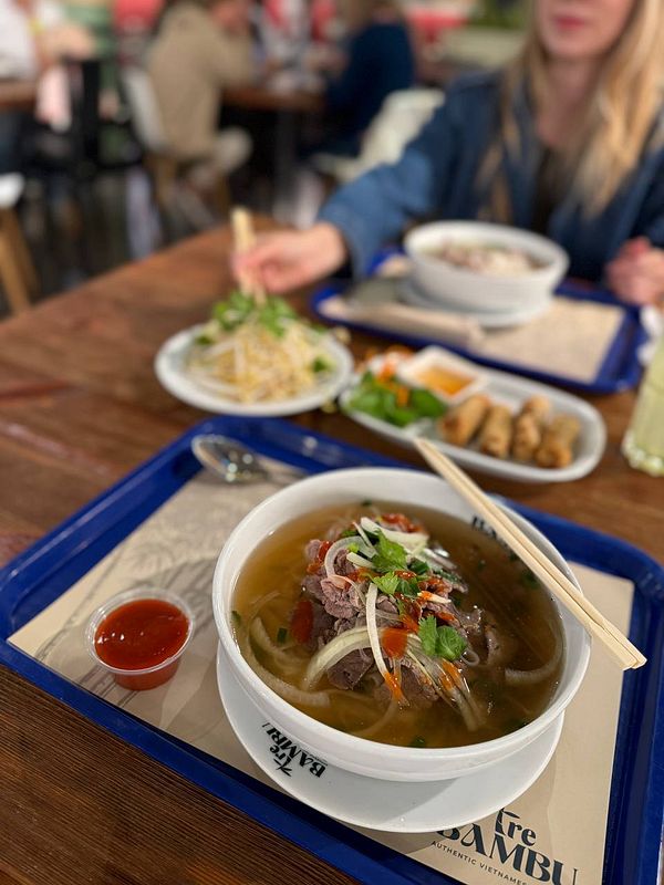 A bowl of Pho is prominently displayed on a tray alongside various side dishes in a casual dining setting.