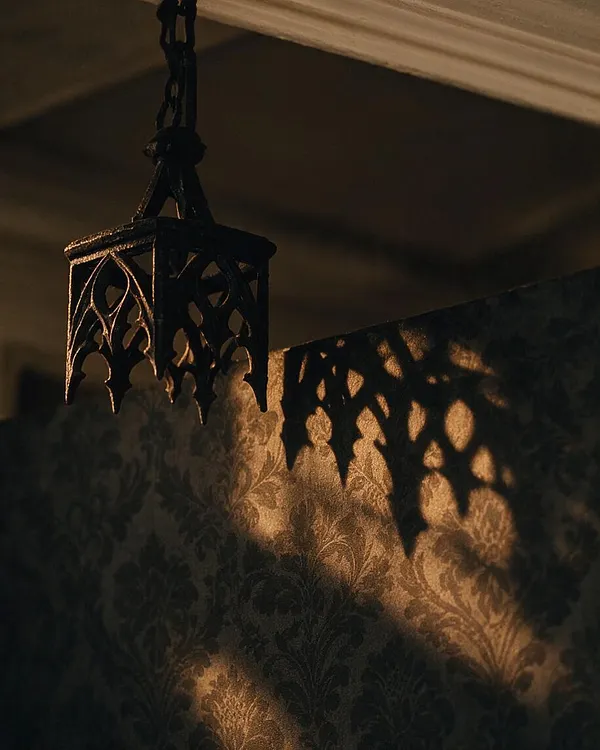 A dark Gothic lantern hangs from the ceiling, made of black metal with pointed arch cutouts and tracery-like detailing. Warm light filters through it, casting intricate shadows across a patterned wallpaper wall in deep green and brown tones.