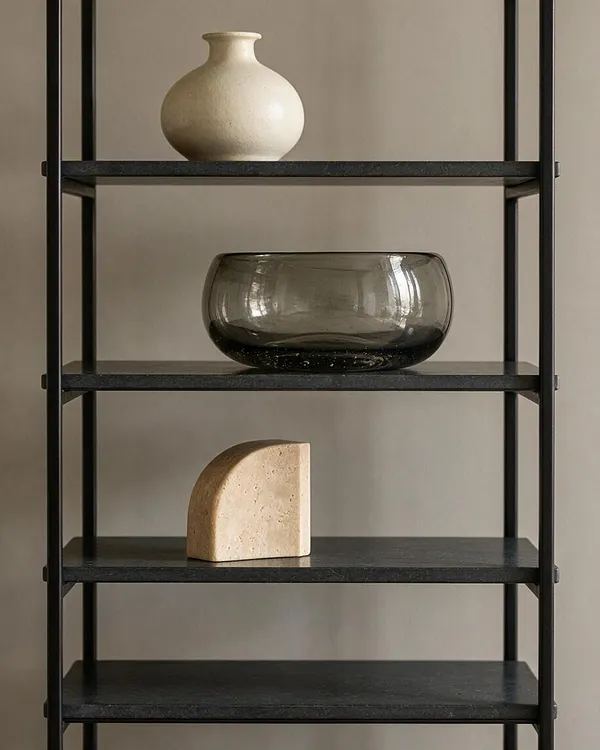 A modern black metal shelving unit against a warm beige wall holds three sculptural decorative objects: a cream ceramic vase on the top shelf, a smoked glass bowl on the middle shelf, and a small stone-like abstract form on a lower shelf. The composition is minimal and balanced with soft natural light.