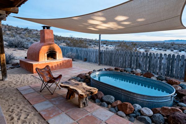 A Southwestern patio set in a desert landscape features a terracotta kiva-style outdoor fireplace beside a square clay-tile terrace on sandy ground. An oval galvanized steel stock tank pool, edged with mixed river stones, sits near a weathered wood picket fence. A leather butterfly chair and a live-edge wood slab table rest under a large beige shade sail, with open views to scrub and distant mountains; warm terracotta contrasts with cool steel and blue water.
