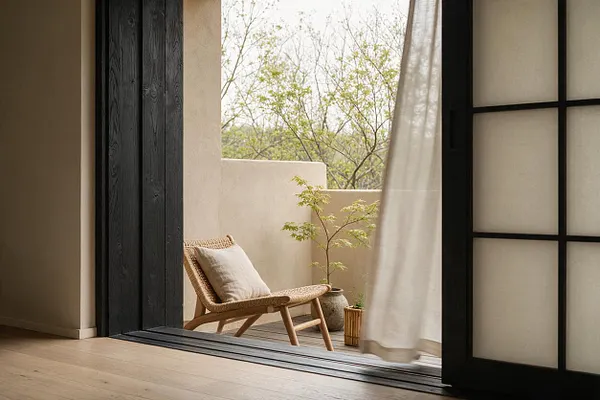 A calm Japandi-style interior opening to a small balcony with a woven lounge chair and light cushion. The scene includes beige stucco walls, dark wood sliding doors, sheer curtain, potted greenery, and warm wood flooring with soft natural daylight.
