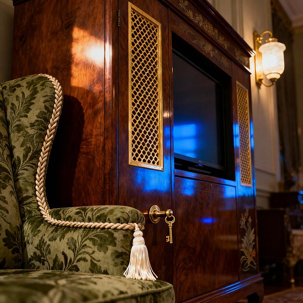 Close-up of a traditional media room shows a high-gloss mahogany TV cabinet with gold lattice grille panels, a brass lock with hanging key, and floral detailing at the base. An adjacent wingback chair is upholstered in olive green damask trimmed with a braided cream cord and tassel, while cream paneled walls and a brass-and-glass sconce cast warm light across the rich wood and deep green palette.