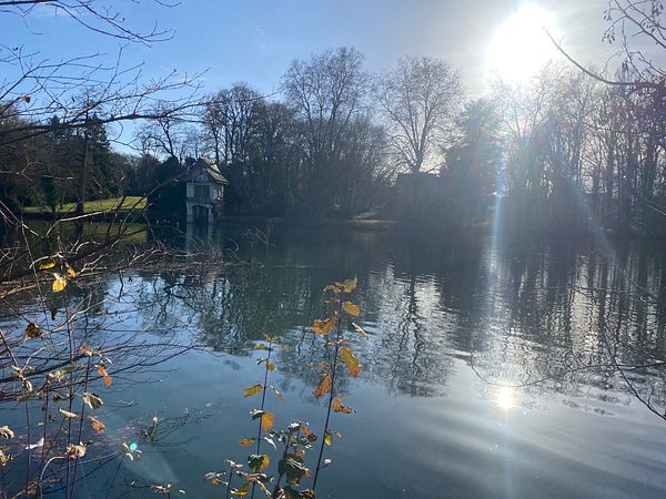 A serene lakeside scene with a small building and trees reflecting in the water under a bright sun.