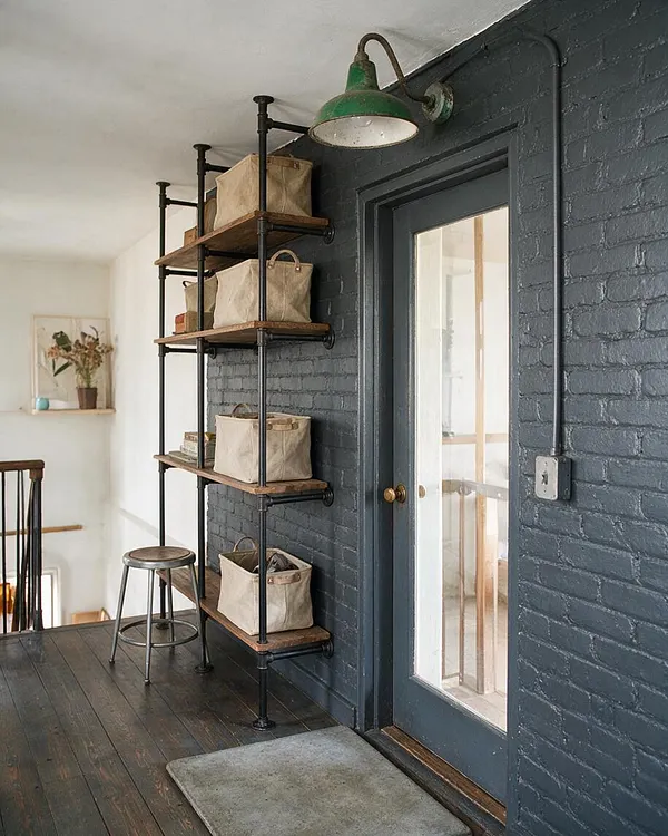 A front porch styled in industrial fashion with dark painted brick walls, a charcoal door with a large glass panel, and exposed black pipe shelving holding beige canvas storage baskets. A green metal wall sconce, wood floorboards, a metal stool, and a simple gray doormat add utilitarian charm.