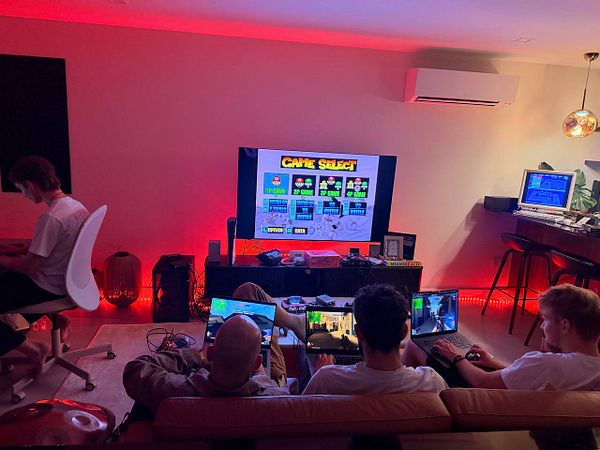 A group of four young men engaging in a gaming session in a cozy room with a large TV, laptops, and retro gaming equipment, illuminated by ambient red lighting.