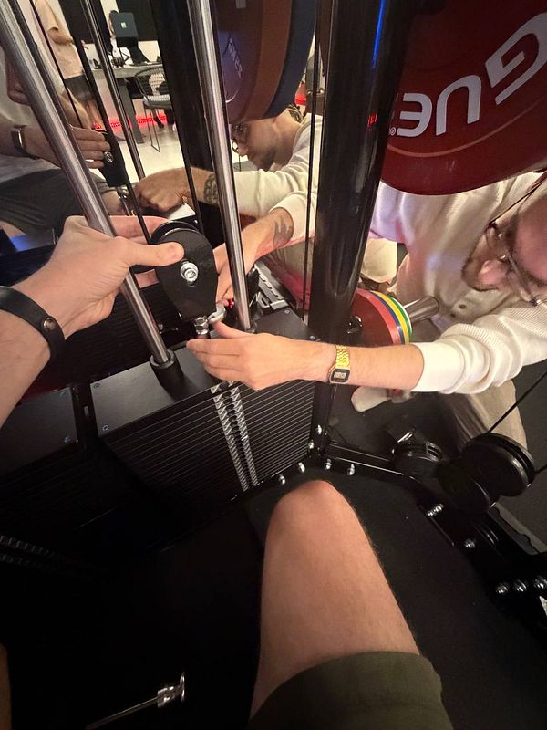 Several people adjust the weight stack on a gym cable machine, with one person's leg visible in the foreground.