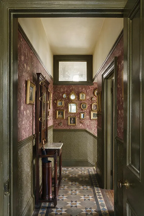 A narrow Victorian entryway with dark green wood trim, burgundy floral wallpaper, and green embossed wainscoting. The patterned tile floor leads to a gallery wall of small gold-framed portraits, with a small marble-topped table and umbrella stand along one side.