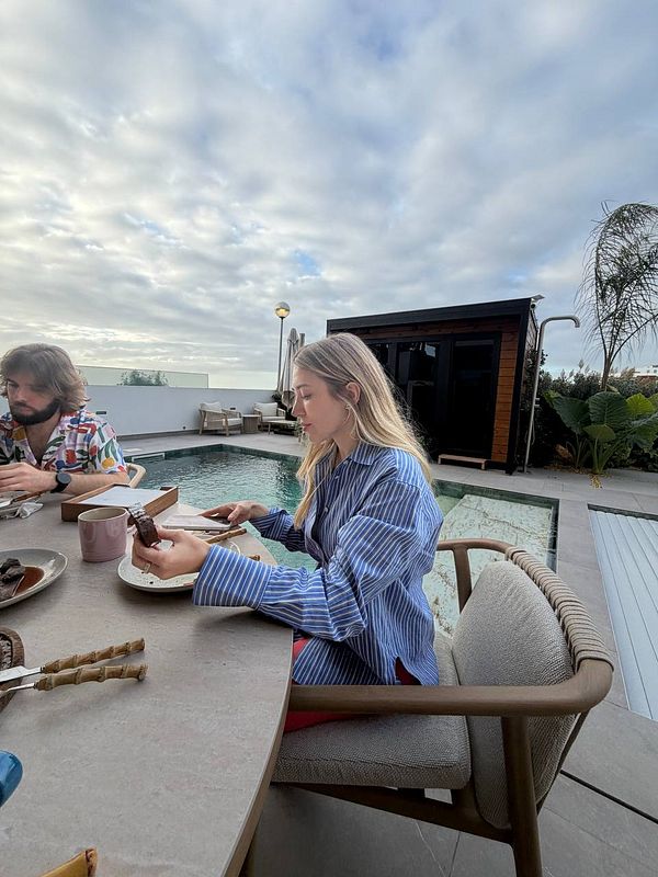 Two people sitting at a table by a pool outdoors with food and drinks, under a cloudy sky.