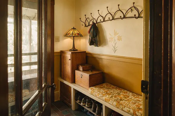 A narrow mudroom with warm mustard walls, a white built-in bench, and a floral patterned cushion above open shoe storage. A dark metal coat rack with curved Art Nouveau lines hangs on the wall above a small wooden cabinet and a stained-glass lamp, while a painted floral motif adds to the decorative style.