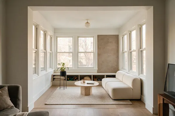 A bright modern sunroom with white-trimmed windows on three sides, a curved cream sofa, a round stone coffee table, and a light textured rug. Built-in storage runs below the back windows, with a small potted plant adding greenery and warm beige walls softening the space.