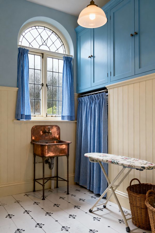 A compact shabby chic laundry room with sky-blue Shaker cabinets, cream beadboard walls, and a white floor patterned with small black motifs. An arched leaded-glass window dressed in blue gingham curtains brings light over a vintage copper wall-mounted sink with exposed plumbing. A curtained utility niche, floral ironing board, wicker laundry baskets, and a milk-glass pendant complete the cozy, nostalgic space.
