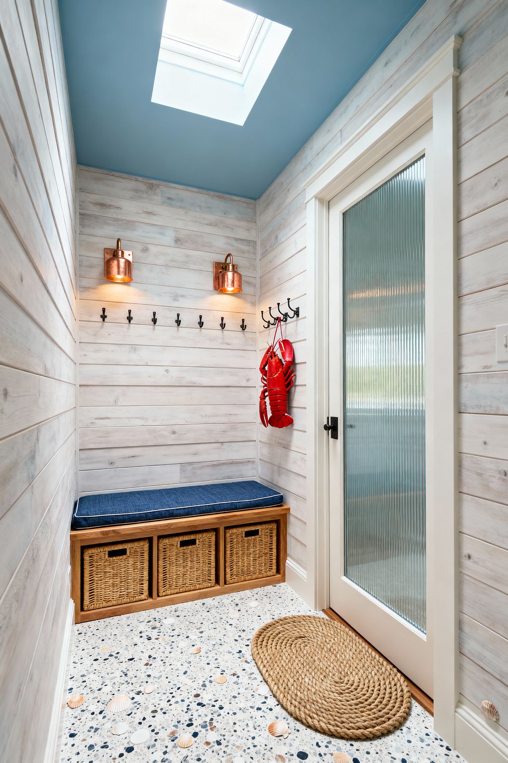 A compact coastal mudroom clad in whitewashed shiplap features a sky-blue ceiling with a large skylight that floods the space with daylight. A built-in wood bench with a navy cushion sits over three rattan baskets, with a row of matte black hooks and two copper-and-glass sconces above; a fluted-glass door with black hardware opens beside a rope doormat, while speckled terrazzo flooring with seashell inlays and a red lobster decoration complete the nautical look.