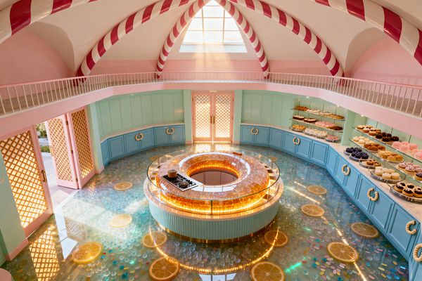 Whimsical pastel kitchen with a circular donut-shaped island lit by amber LEDs and a built-in cooktop, surrounded by mint cabinets. Glass shelves display pastries above pale stone counters. The glossy aqua epoxy floor embeds lemon slices and confetti shapes. A vaulted ceiling with red-and-white candy-cane ribs crowns the room, with daylight through an arched window and pink lattice double doors.