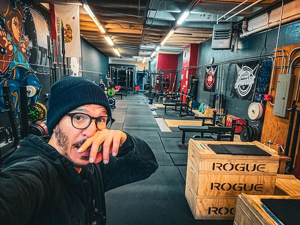 Photograph of a gym weightlifting area focusing on a squat rack with a loaded barbell and exercise mats.