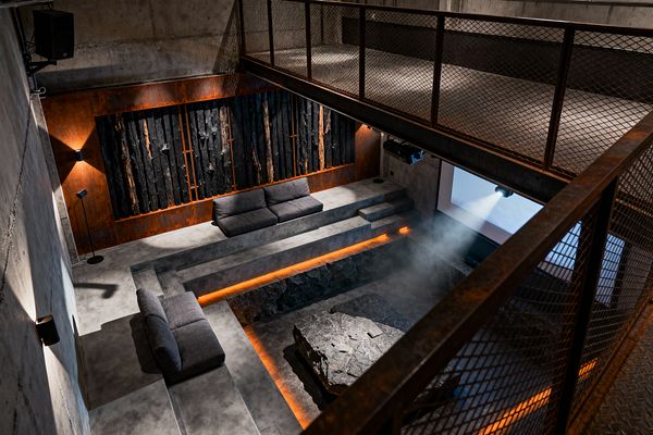 A double-height industrial media room with raw concrete walls, a sunken seating pit, and tiered concrete platforms with low gray sofas. A projector beams onto a wall screen; amber LED strips trace the pit’s edge around a rough-hewn stone table. A backdrop of Corten steel holds a charred-wood art panel, while a mesh metal mezzanine overlooks the space and black wall lights add focused illumination.