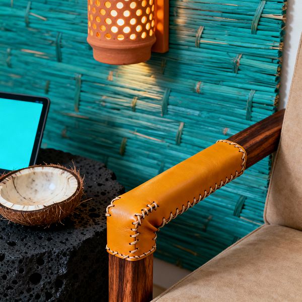 Close-up of a tropical media room corner with a teal woven reed wall panel, a perforated terracotta sconce casting warm light, and a chair in beige suede with a striped hardwood frame wrapped in tan leather stitching at the arm. A porous black lava-stone side table holds a coconut-shell bowl beside the edge of a tablet, combining natural textures and earthy tones to create a relaxed, island-inspired setting for casual screen time.