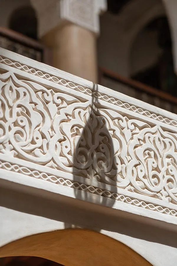 A close-up of an ornate white carved railing with swirling floral arabesque patterns and delicate lattice borders. Soft shadows fall across the textured surface, with blurred arches and a warm beige wall in the background.
