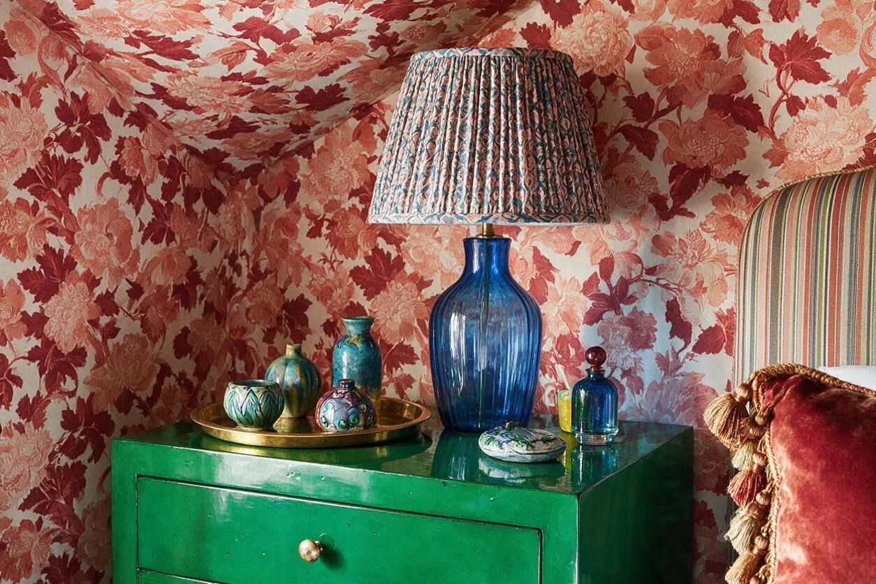 A maximalist corner vignette with pink and coral floral wallpaper covering the walls and sloped ceiling. A glossy green nightstand holds a tall cobalt-blue glass lamp, a brass tray with small decorative vases, and a few colorful glass accessories beside a striped upholstered headboard and a red velvet tassel pillow.