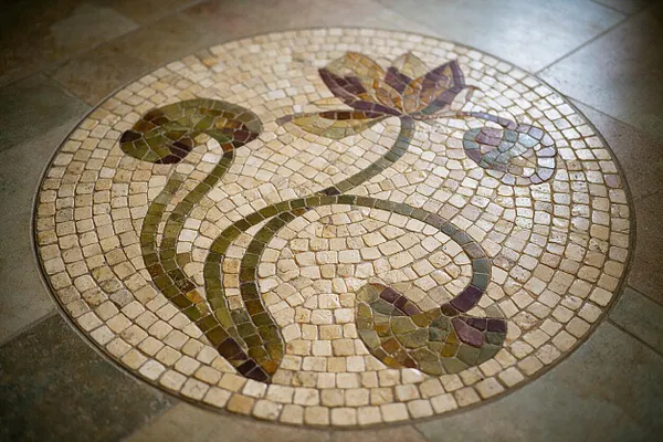 A circular floor mosaic set into stone tile, featuring a flowing Art Nouveau floral design with curving green stems and stylized blossoms in cream, olive, purple, and blue glass-like pieces. The surrounding floor is made of large neutral stone tiles.