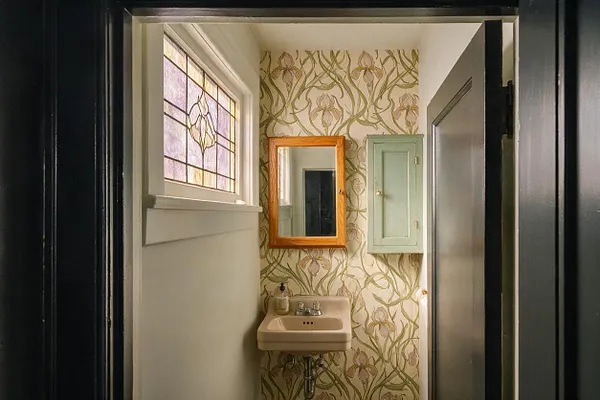 A narrow powder room with Art Nouveau floral wallpaper, a small beige wall-mounted sink, and a wood-framed mirror above it. A stained-glass window on the left adds soft colored light, while a pale mint medicine cabinet and dark doorway trim complete the vintage look.