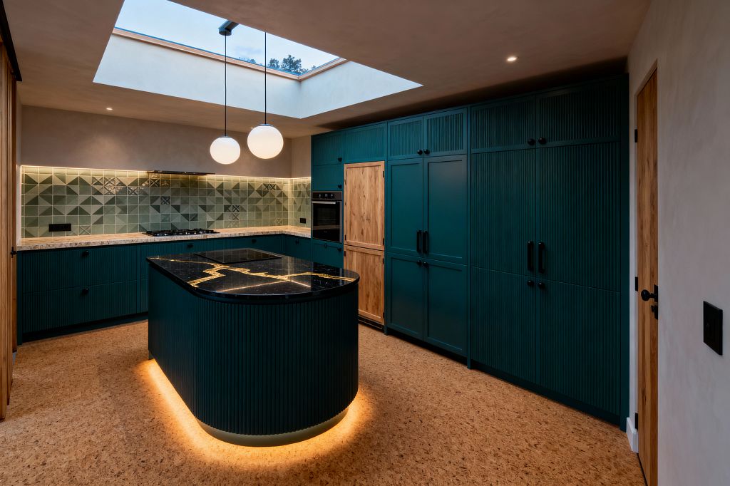 A contemporary kitchen with deep teal, vertically reeded cabinets and a curved island beneath a large skylight. The island has a black stone countertop with bold gold veining and warm LED base lighting. A sage green geometric tile backsplash runs along the perimeter above light speckled stone counters, with matte black hardware and appliances. The space features cork flooring, two globe pendants, and pale plaster walls for a warm, modern look.