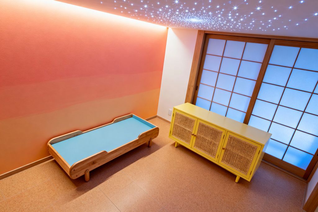 A modern children’s room features a peach-to-coral gradient wall lit by concealed cove lighting, a low light-wood toddler bed with a sky-blue mattress, and a canary-yellow cabinet with woven cane doors. Shoji-style sliding doors in a natural wood frame diffuse cool blue light, while a starry fiber‑optic ceiling sparkles above warm cork flooring for a clean, playful atmosphere.