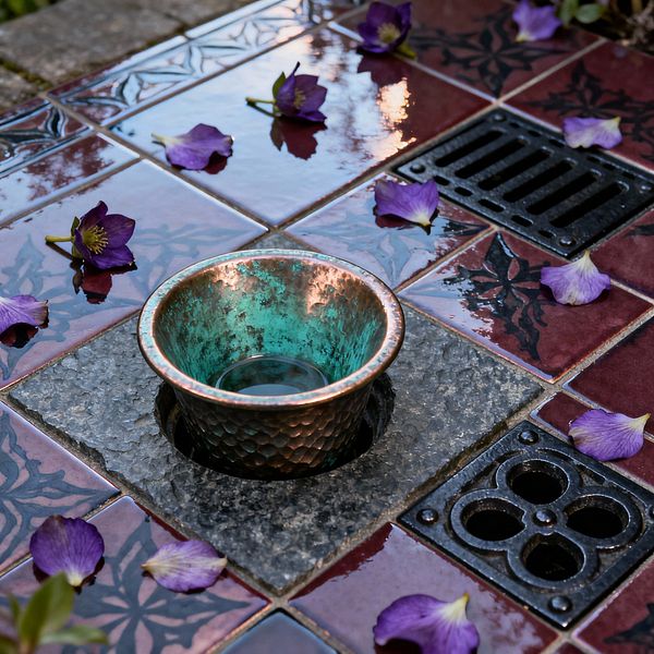 Close-up of a patio shows glossy oxblood glazed tiles patterned with black leaf tracery. A rough-cut square of gray stone frames a recessed opening holding a small hammered copper cup with a green verdigris interior. Nearby are two cast-iron drains, one rectangular and one with a quatrefoil design. Purple flower blossoms and petals lie across the wet surface, reflecting light and creating a moody, Gothic-inspired outdoor scene.