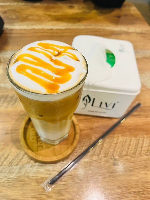 A glass of coffee topped with cream and caramel drizzle is presented on a wooden table.