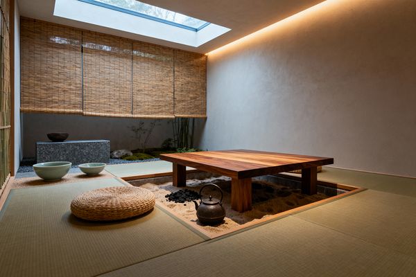 A minimalist Japanese dining room features a low solid-wood table set over a sunken sand hearth with charcoal and a cast-iron kettle, surrounded by tatami mats and round woven floor cushions. A skylight and warm cove lighting wash plaster walls, while bamboo roll-up blinds filter views to an indoor rock garden with moss, pebbles, and a granite bench; simple ceramic bowls rest on a straw mat.