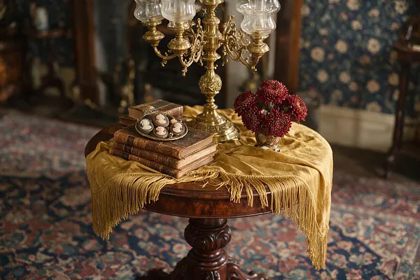 A Victorian-inspired round wooden side table is draped with mustard-gold fabric and styled with stacked antique books, a brass candelabra, a small bouquet of dark red flowers, and cameo jewelry. The background shows a richly patterned rug and floral wallpaper, creating a formal old-world atmosphere.