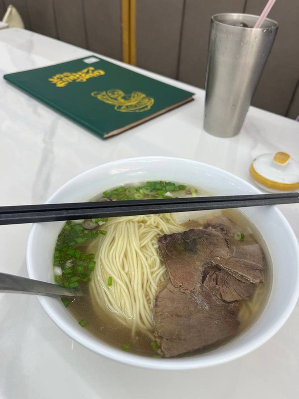A bowl of noodle soup with slices of beef, chopped green onions, and clear broth, with black chopsticks resting on the bowl.