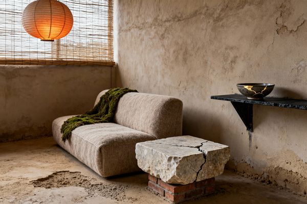 A Wabi-Sabi living room shows a low, armless sofa in soft taupe wool against rough, limewashed plaster walls. A moss-green throw lies on the seat, a cracked stone slab set on red bricks serves as a table, and a slim black shelf holds a ceramic bowl mended with gold kintsugi. Bamboo blinds filter daylight while a round paper lantern glows warmly above an uneven concrete floor.