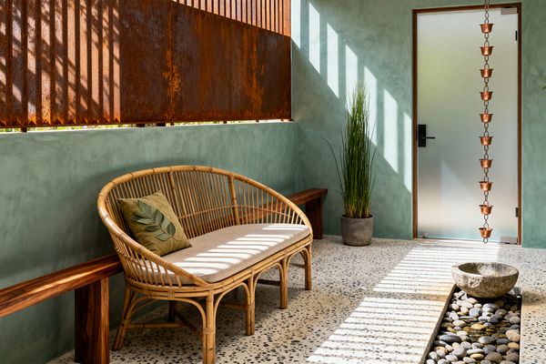 Biophilic mudroom with sage-green plaster walls and speckled terrazzo floor. A rattan settee with beige cushion and leaf pillow faces a live-edge wooden bench. A pebble-lined water rill with a carved stone bowl runs along the floor beneath a copper rain chain by a frosted glass door. Corten steel slats filter sunlight, casting striped shadows. A tall potted grass adds greenery.