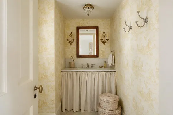 A narrow powder room styled in French Country fashion with pale yellow toile-like wallpaper, a dark wood framed mirror, and antique brass wall sconces. A small vanity with a cream skirted curtain, brass faucet, and woven baskets creates a soft, charming, old-world look.