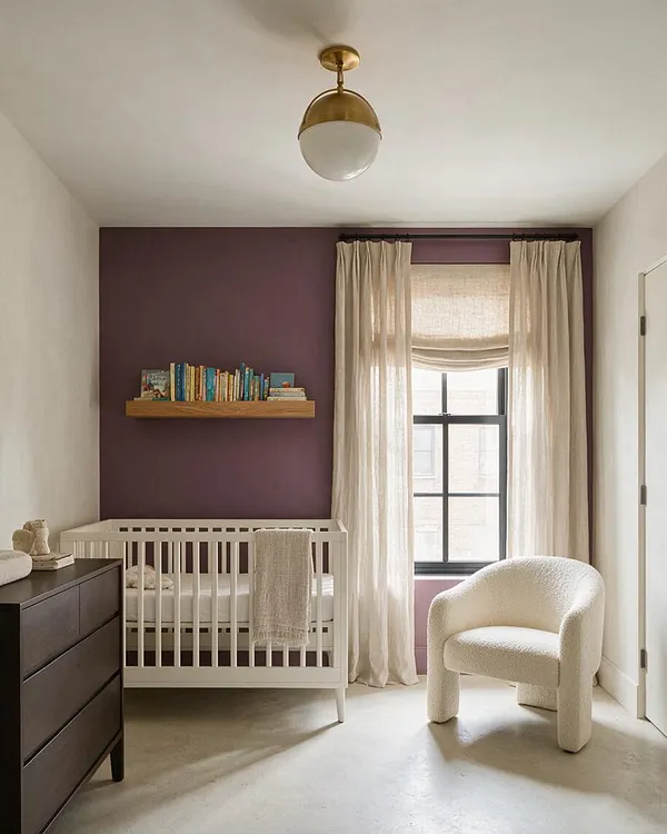A modern nursery with a deep plum accent wall, a white crib, and a floating wood book ledge above it. Cream curtains frame a window, while a textured ivory chair and dark dresser add soft contrast under a brass ceiling light.