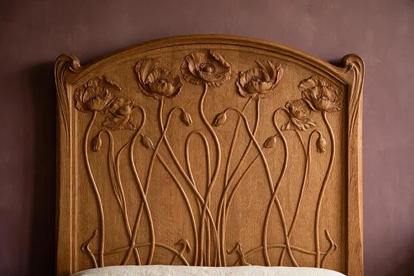 A carved wooden headboard with an arched top and flowing Art Nouveau floral relief, featuring tall poppy-like stems and blossoms across the upper panel. It stands against a muted mauve wall, with the edge of a light-colored bed covering visible at the bottom.
