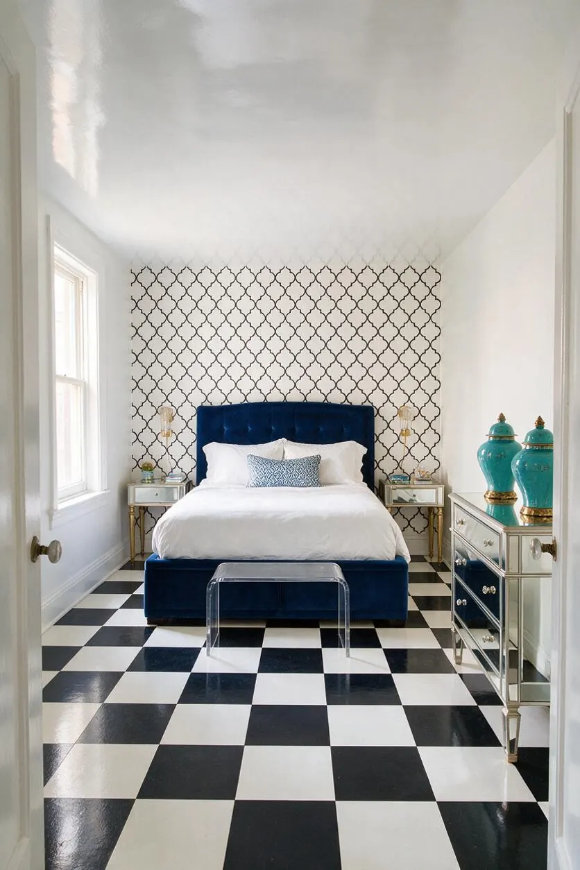 A bright master bedroom with a glossy black-and-white checkerboard floor, white walls, and a patterned accent wall behind a deep navy velvet bed. Matching mirrored nightstands with gold trim and turquoise decorative jars add Hollywood Regency glamour, while a clear acrylic bench sits at the foot of the bed.