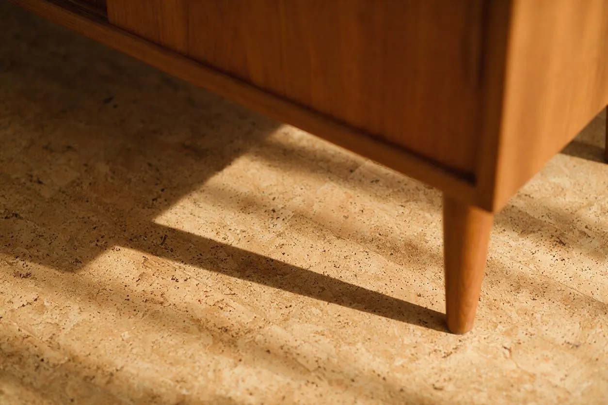 A close-up of a walnut mid-century modern cabinet with tapered legs casting long shadows onto a light travertine floor. The stone surface shows natural speckling and soft beige tones, creating a warm, retro-inspired interior detail.
