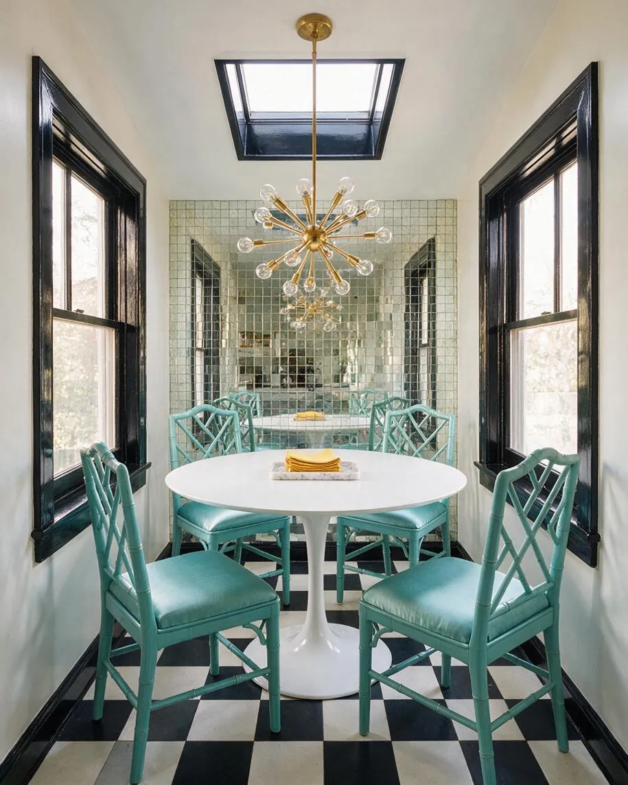 A narrow sunroom with black-trimmed windows, a black-framed skylight, and a round white pedestal table surrounded by four turquoise bamboo-style chairs. A mirrored mosaic wall reflects a gold starburst chandelier, and the room has a black-and-white checkered floor.
