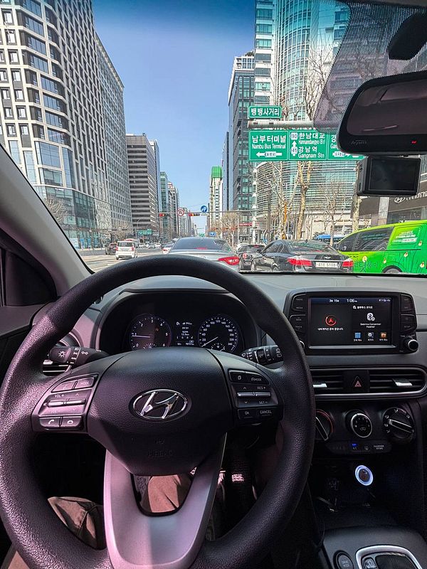 A view from inside a car driving through Gangnam, showcasing the urban landscape and dashboard.